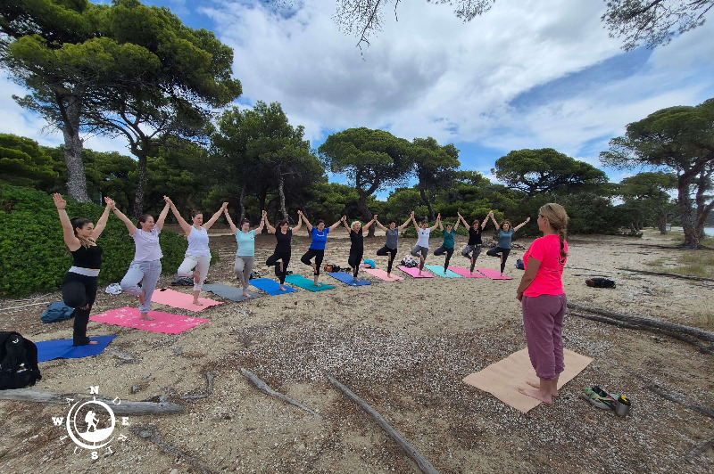 Εκπαιδεύτρια καθοδηγεί ομάδα σε στάση yoga με τα χέρια ψηλά, κοντά στη θάλασσα στον Σχινιά